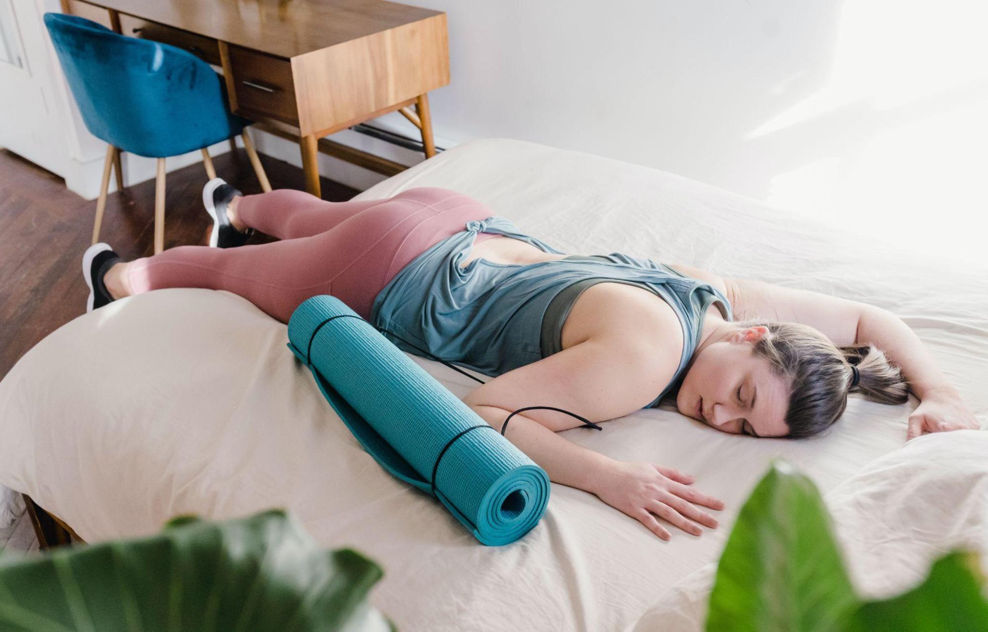 woman in athletic attire resting on a bed with a yoga mat